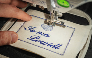 Close-up of an embroidery machine embroidering a German saying on white linen fabric, held in an embroidery frame.