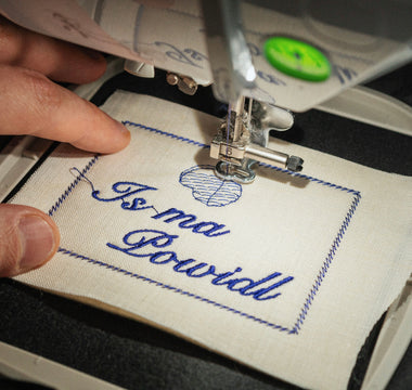 Close-up of an embroidery machine embroidering a German saying on white linen fabric, held in an embroidery frame.