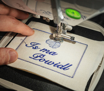 Close-up of an embroidery machine embroidering a German saying on white linen fabric, held in an embroidery frame.