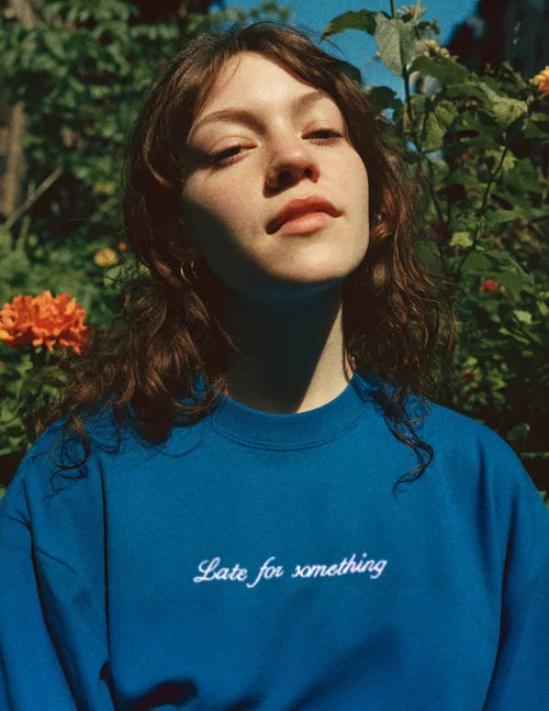 Blaues besticktes Sweatshirt mit weißem Schriftzug auf der Brust, getragen von Model im Garten bei natürlichem Licht